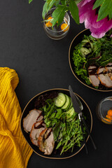 Hearty delicious lunch, fresh salad with cucumbers and microgreens of peas and with stewed pork, top view of the dark table with food and a vase of flowers
