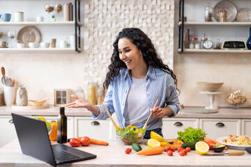 Obraz na płótnie Canvas Online communication concept. Happy latin woman preparing salad and talking on video chat via laptop in kitchen