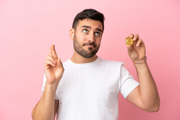 Young man holding a Bitcoin isolated on pink background with fingers crossing and wishing the best