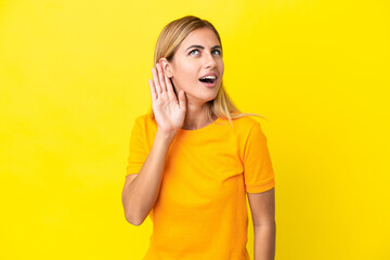 Blonde Uruguayan girl isolated on yellow background listening to something by putting hand on the...