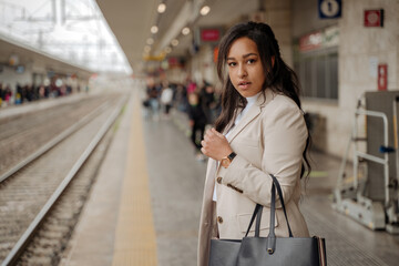 Portrait of beautiful successful business woman looking at camera standing at railway station, copy space. Pensive tourist holding bag  waiting for train. Transportation, travel concept