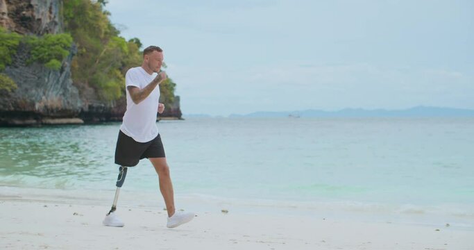 Young caucasian man with artificial bionic legs prosthesis running and jogging on a tropical beach in a sunny day. Healthy and active disability sportsman training exercise workout in morning seaside.