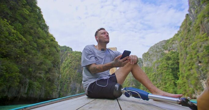 Man with artificial bionic leg prosthesis sitting on the front of long tail boat listening music while enjoying amazing beautiful seascape during summer vacation travel in Phi Phi Islands, Thailand.