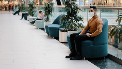 Obraz premium Young indian guy in medical mask protecting health during pandemic sitting in armchair indoors in shopping center mall resting relaxing nervous anxious waiting for appointment long expectation concept