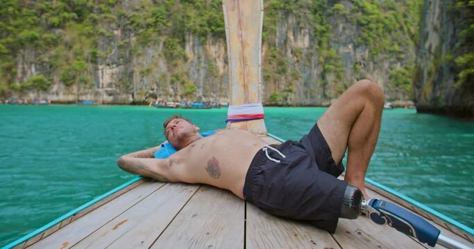 Man with artificial bionic legs prosthesis relaxing by laying down and sleeping on the front of long tail boat enjoy beautiful seascape during summer vacation travel in Phi Phi Islands, Thailand.