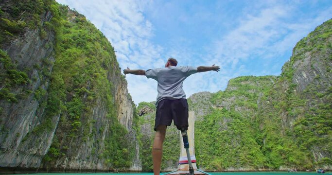 Male with artificial bionic leg prosthesis standing on the front of long tail boat seeing an amazing beautiful seascape and raise his hand with a smile during summer vacation travel. Phi Phi, Thailand