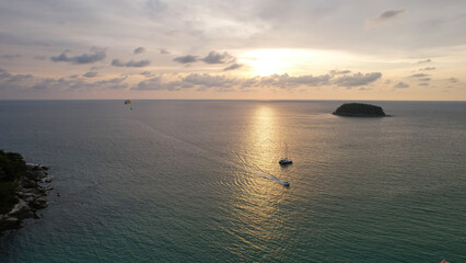 Parasailing at sunset with a view of the island. Parachute flight in the midst of a colorful sunset. Small waves, people relax on the beach, palm trees grow, there are hotels. Boats float. Asia Phuket