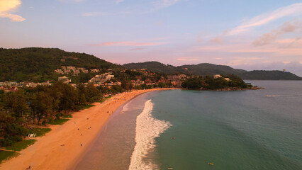 Orange sunset with a view of the beach. People are resting, seeing off the sun over the horizon. Small waves. Clean sand. Hills, clouds and the city are visible in the distance. Dogs are running. Kata