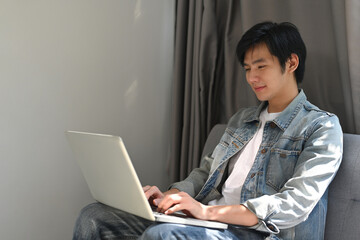 Portrait of smiling Asian Man Sitting on the sofa and working on PC laptop in the living room, Using computer for study or education.