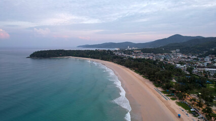 Fototapeta na wymiar Early dawn on the island of Phuket. Top view from the drone on the sand and waves. People are walking along the beach. Calm environment. Green hills and clouds in sky. Lights of the city are shining