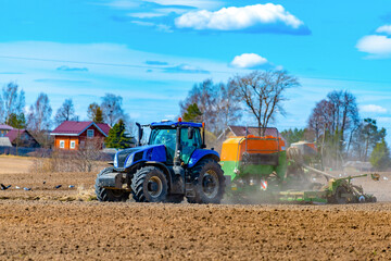 A tractor with a seeder sows grains of wheat or oats in the spring in a field for growing grain products against the backdrop of village houses.