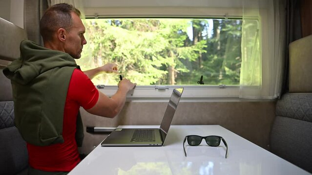 Caucasian Men in His 40s Working on His Laptop In His Camper Dinette Area and Opening Side Window. Travel and Working Theme.