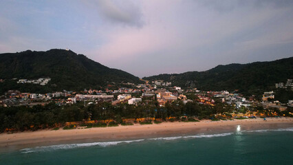 View of the beach and the island from a height. Sunset. People walk on the beach, relax. There are clouds in the sky. Green hills on the island of Phuket. There are hotels, lanterns are lit. Journey