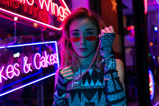 Millennial enigmatic pretty girl with unusual dyed hairstyle near glowing neon wall at night. Blue hair, golden sequins as freckles,nose piercing. Mysterious hipster teenager in glasses
