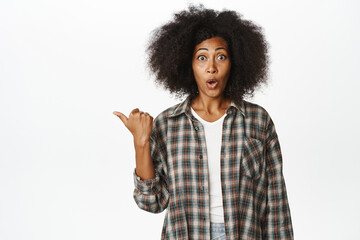 Surprised african girl points left with amazed gasp and disbelief, asking question about store advertisement, intrigued with promo offer, white background