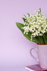 A bouquet of lilies of the valley stands in a mug on a pink notebook