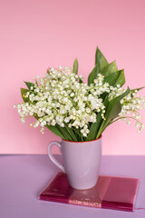 A bouquet of lilies of the valley stands in a mug on a pink notebook