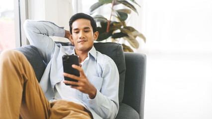 Young adult southeast asian man relax on sofa using smartphone for online leisure at home background with copy space