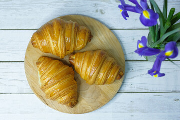 Fresh baked croissants on wooden cutting board on white wooden table with flowers Irises. Flat lay.