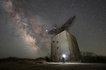 A person exploring an abandoned military base coming across a radar tower at night under the Milky Way Galaxy 
