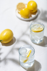 Two glass of water with ice cubes and lemon slices and a plate of lemons stand on a white table. Top view