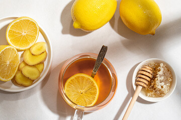 A mug of tea with lemons and a ginger root and little plate of honey with a wooden spoon on a table covered with a white tablecloth. Folk way of treating colds