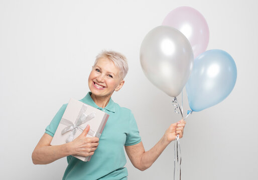 Amazed elderly woman with short hair holding balloons and gift boxes over white gray background.