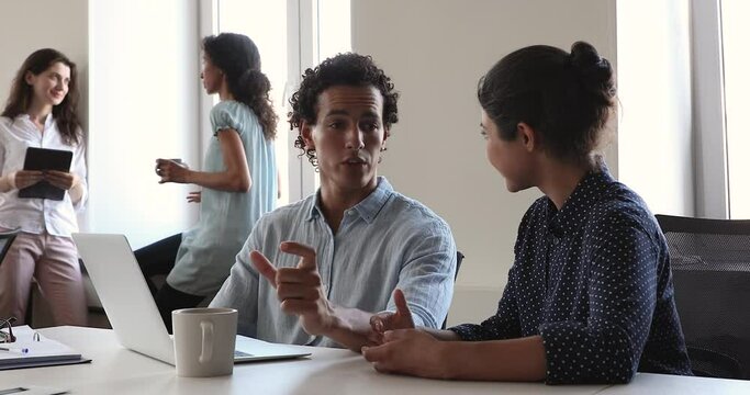 Professional Hispanic male tutor teach young Indian lady new staff member explain work with documents in corporate network app at cozy office space. Multiethnic coworkers engaged in teamwork by laptop