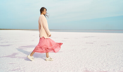 Cheerful walking woman in oversize sweater and blowing pink dress at paradise white coast of pink...