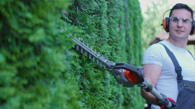Handsome caucasian man in overalls, protective glasses and gloves pruning hedge with electric trimmer. Competent gardener enjoying seasonal work with plants outdoors. Concept of trimming.