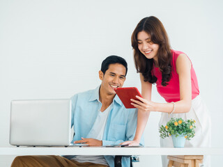 Happy smiling Asian business couple, young man and woman, colleagues using tablet and laptop computer on table, looking at screen, working together at the white clean small office background.