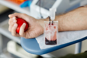 Close up of male hand squeezing stress ball while donating blood at medical volunteer center focus on blood bag in foreground, copy space