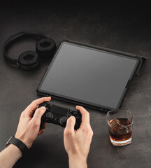 Gaming process with joystick in hand. Mockup template on dark structural background. Gamer workplace with headphones, tablet and whiskey glass with ice.
