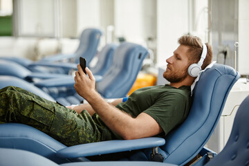 Side view portrait of man donating blood in comfort and watching videos via smartphone