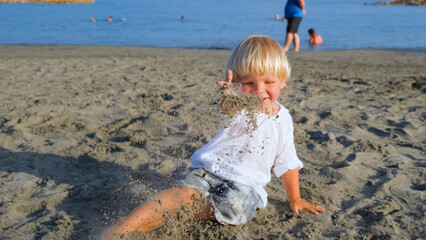 little boy in a white shirt plays on the beach throwing sand at the camera