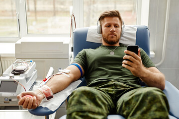 Portrait of young military man donating blood with comfort and watching videos at smartphone, copy...