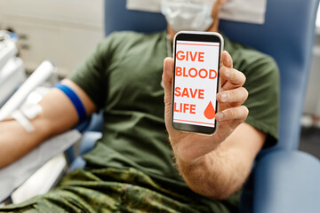 Closeup of soldier donating blood and holding smartphone to camera with Give blood save life on...