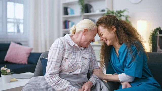 Female nurse keeping company for senior lady at home, women talking and laughing
