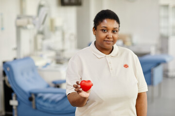 Waist up portrait of young black woman holding red heart at blood donation center and looking at...