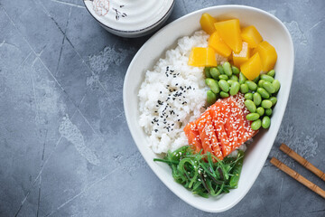 Poke bowl with salmon, edamame beans, mango and seaweed salad, above view on a grey concrete background, horizontal shot with space