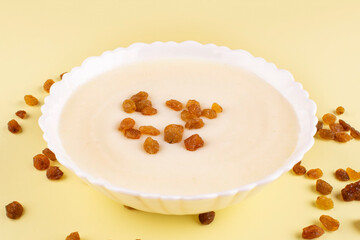 Semolina porridge with raisins in a white plate (bowl) on a yellow background close-up. Side view. Baby food. Contains gluten in the composition.