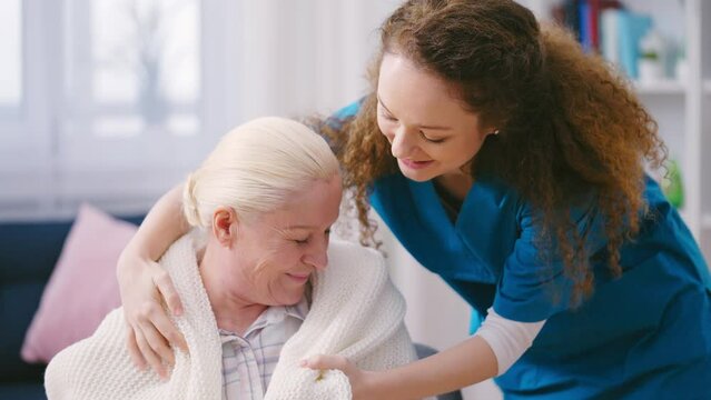 Caring female volunteer covering elderly woman with warm blanket, home care