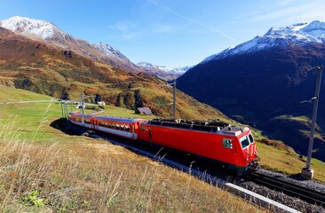 A tourist train traveling on the green grassy hillside on a beautiful autumn day and snowy alpine mountains towering under blue sunny sky in background, in Andermatt, canton of Uri, Switzerland