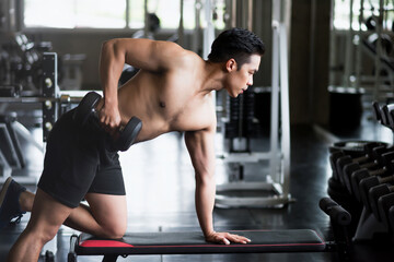 Fototapeta na wymiar Fit Asian young man doing one-arm dumbbell on bench in gym