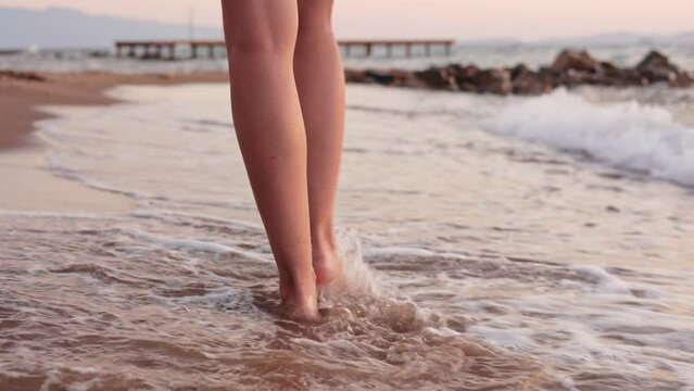 Close up slim female legs and feet walking barefoot on sandy beach at sunset or sunrise. Splashes of water at seaside. Woman tourist on summer vacation.