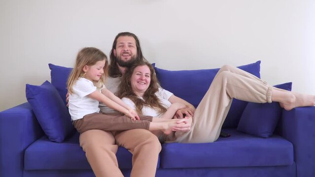 Happy family watching TV on the sofa. Weekends at home on the couch together. Have fun and play. Mom and dad hug their daughter. Girl in a joyful childhood. Cozy house.