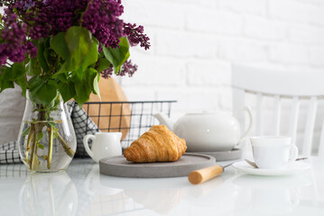 Fresh croissant on a grey plate on a table with a white teapot and lilacs