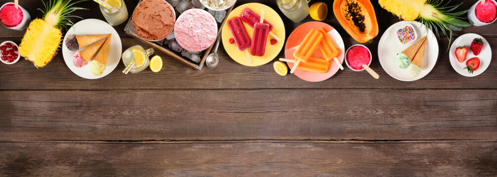 Cool summer foods top border. Selection of ice cream, popsicles and fruit. Above view over a dark wood banner background.