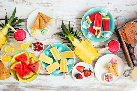 Cool summer foods table scene. Collection of ice cream, popsicles and fruit. Overhead view on a rustic white wood background.