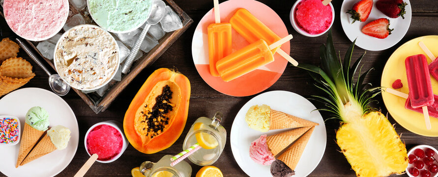 Refreshing summer foods table scene. Assorted ice cream, popsicles and fruit. Top down view over a dark wood banner background.